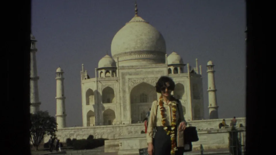 1974 8mm frame of a woman posing under the Taj Mahal dome line, with visitors and landscaped grounds visible behind her.