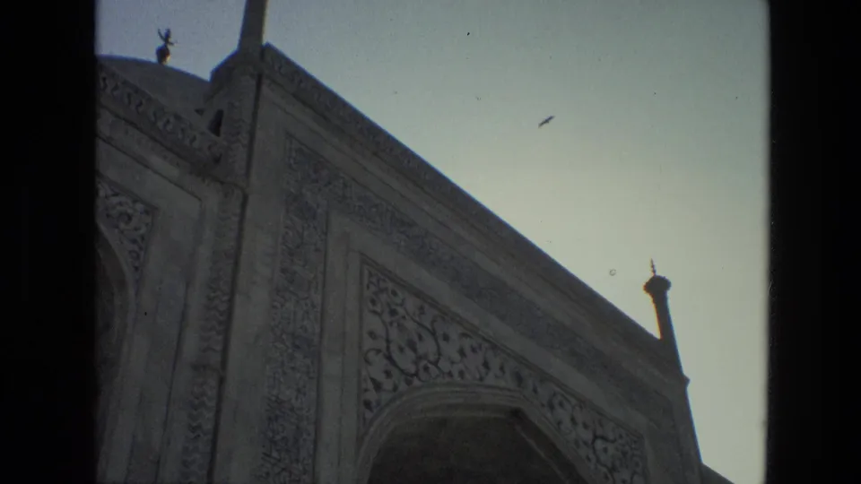1974 India 8mm frame showing an old mosque facade from below, with ornate carving, an arched opening, and a bird overhead.