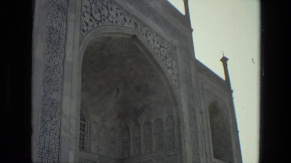 1974 India 8mm frame holding on the old mosque facade, with the arch, decorative carvings, roofline finials, and sky in view.