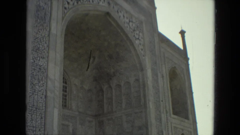 1974 8mm view in India of an ornate arched facade with carved panels and calligraphy, partly obscured by a dark doorway or window edge.
