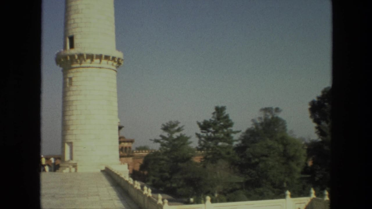 A tall white minaret rises beside marble walkways and trees.