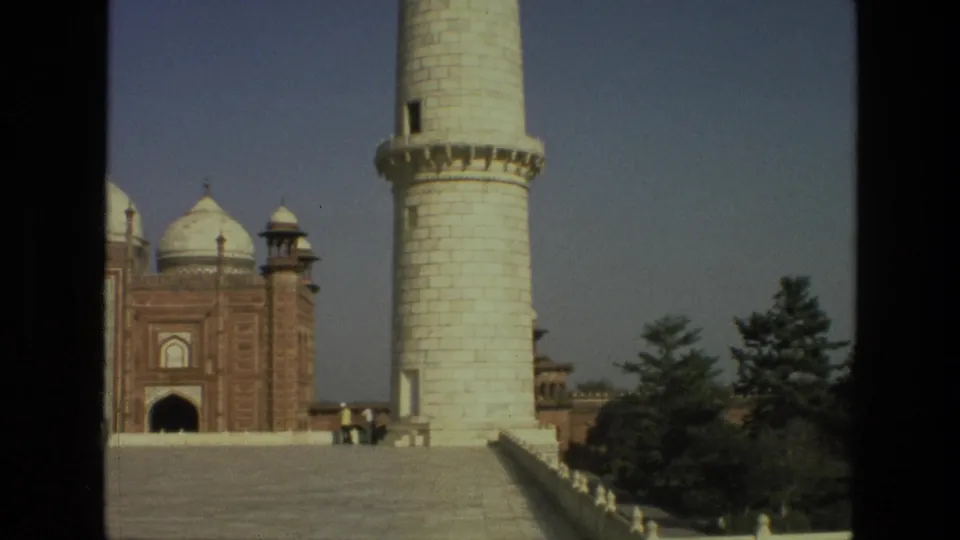 1974 8mm view of a Taj Mahal minaret with red sandstone gateway architecture and small visitors on the platform.