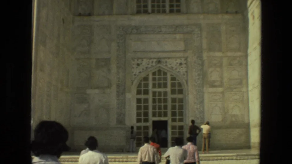 A handheld 8mm view shows tourists gathered before a tall arched doorway and carved facade at an Indian Mughal-style landmark.