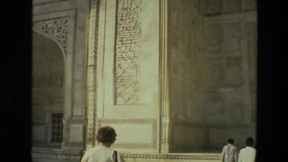 The frame shows the side of a Mughal-style stone facade in India with calligraphy panels and tourists walking along the courtyard edge.