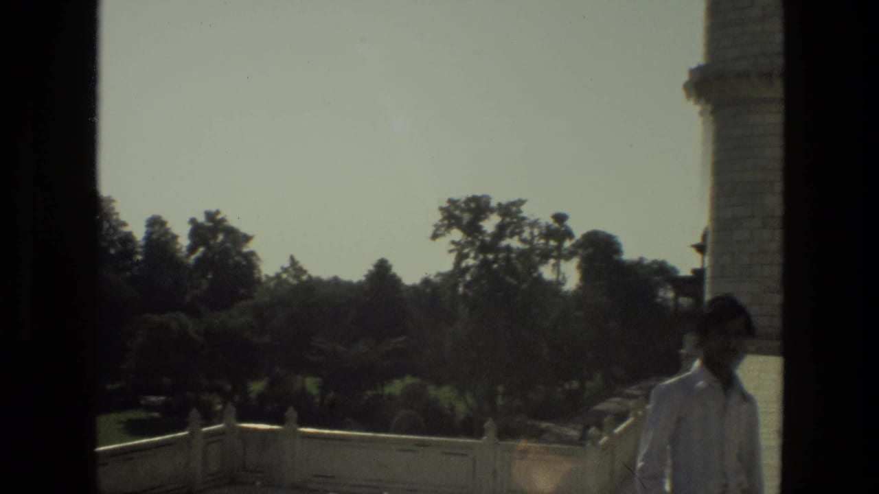 Man in a light shirt standing near a stone tower with gardens behind him.