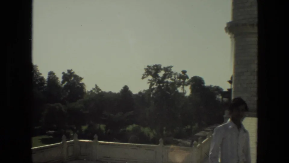 1974 India travel frame with a man in a light shirt near a Taj Mahal minaret edge, with garden trees and railings in the background.