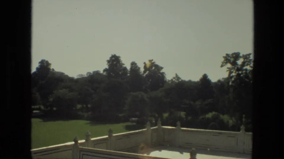 1974 8mm view across the Taj Mahal gardens in India, with trees, lawn, white railing, and a monument edge along the frame.