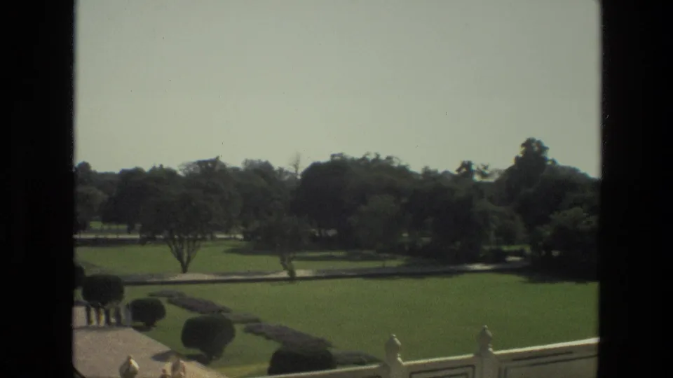 1974 8mm view across Taj Mahal gardens in India, with lawns, hedges, trees, paths, and distant monument architecture.