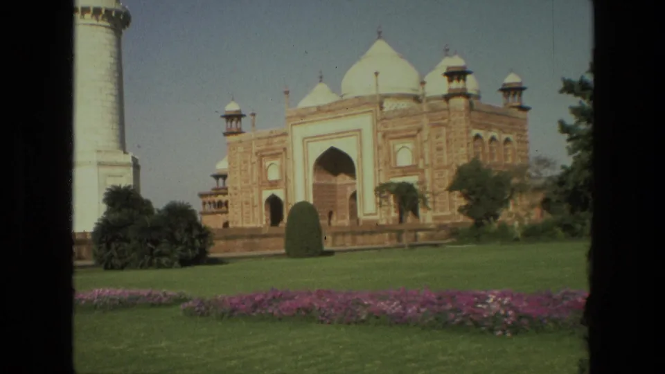 1974 8mm footage frames the Taj Mahal complex in India with a minaret, green lawn, shrubs, and purple flowerbeds.