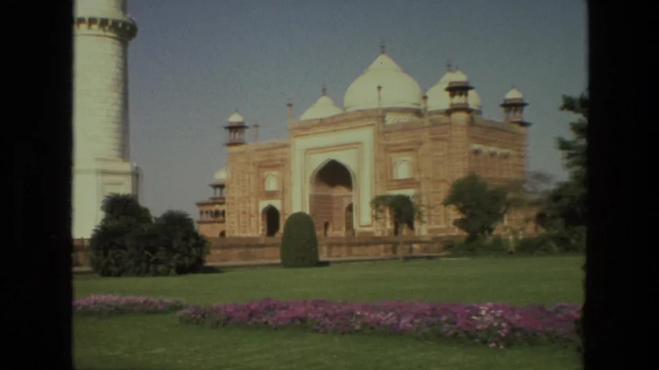 1974 India home-movie view of Taj Mahal grounds with red sandstone architecture, a white minaret, shaped trees, grass, and purple flowers.
