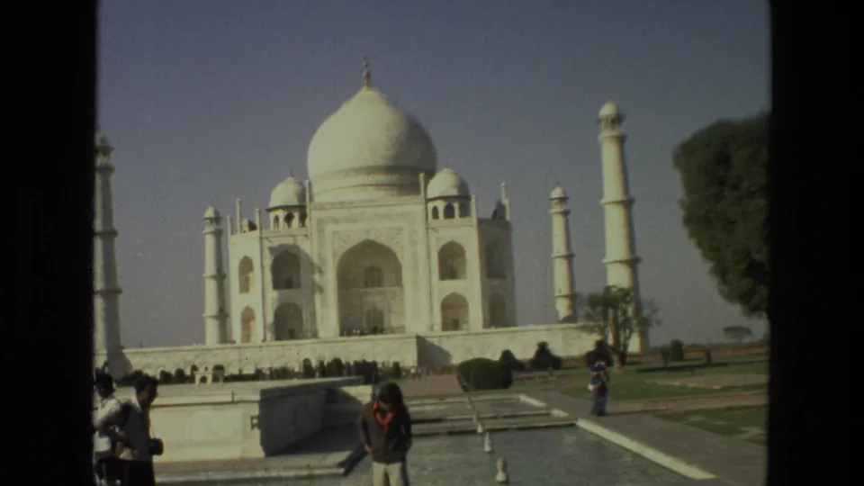 1974 India home-movie frame of the Taj Mahal across the gardens, with visitors in front, minarets, trees, and reflecting-pool borders visible.