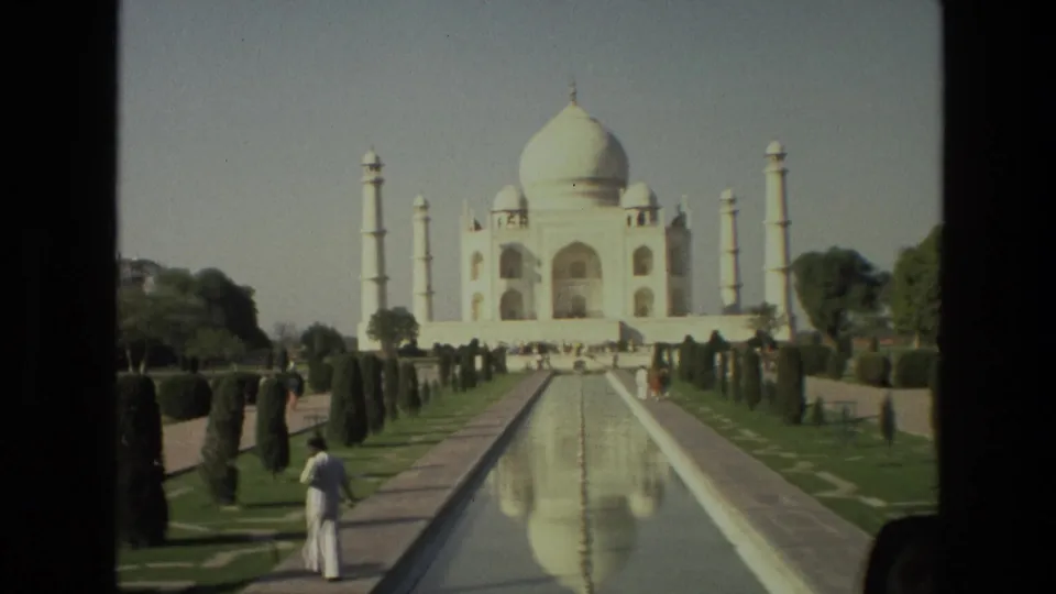 1974 8mm frame looking along the Taj Mahal reflecting pool, with a visitor in white near the foreground and garden plantings on both sides.