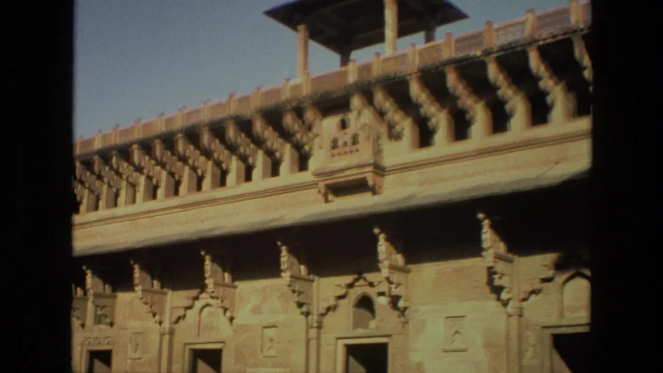 1974 India frame showing sunlit palace-style architecture with carved brackets, balconies, and arched wall details.