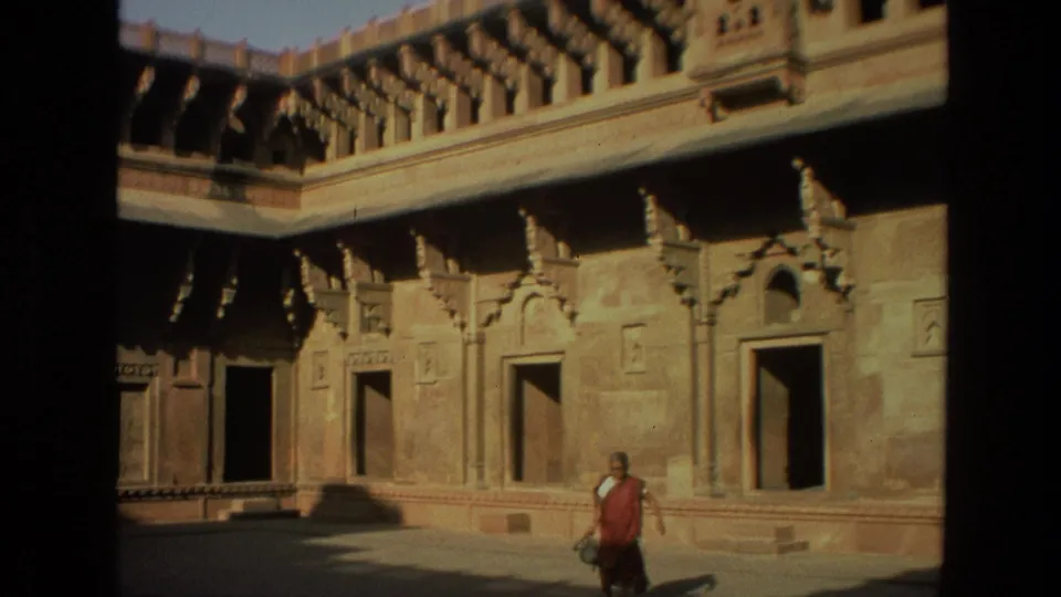 1974 India 8mm frame showing a robed person walking through a sunlit shrine courtyard with carved stone walls and open doorways.