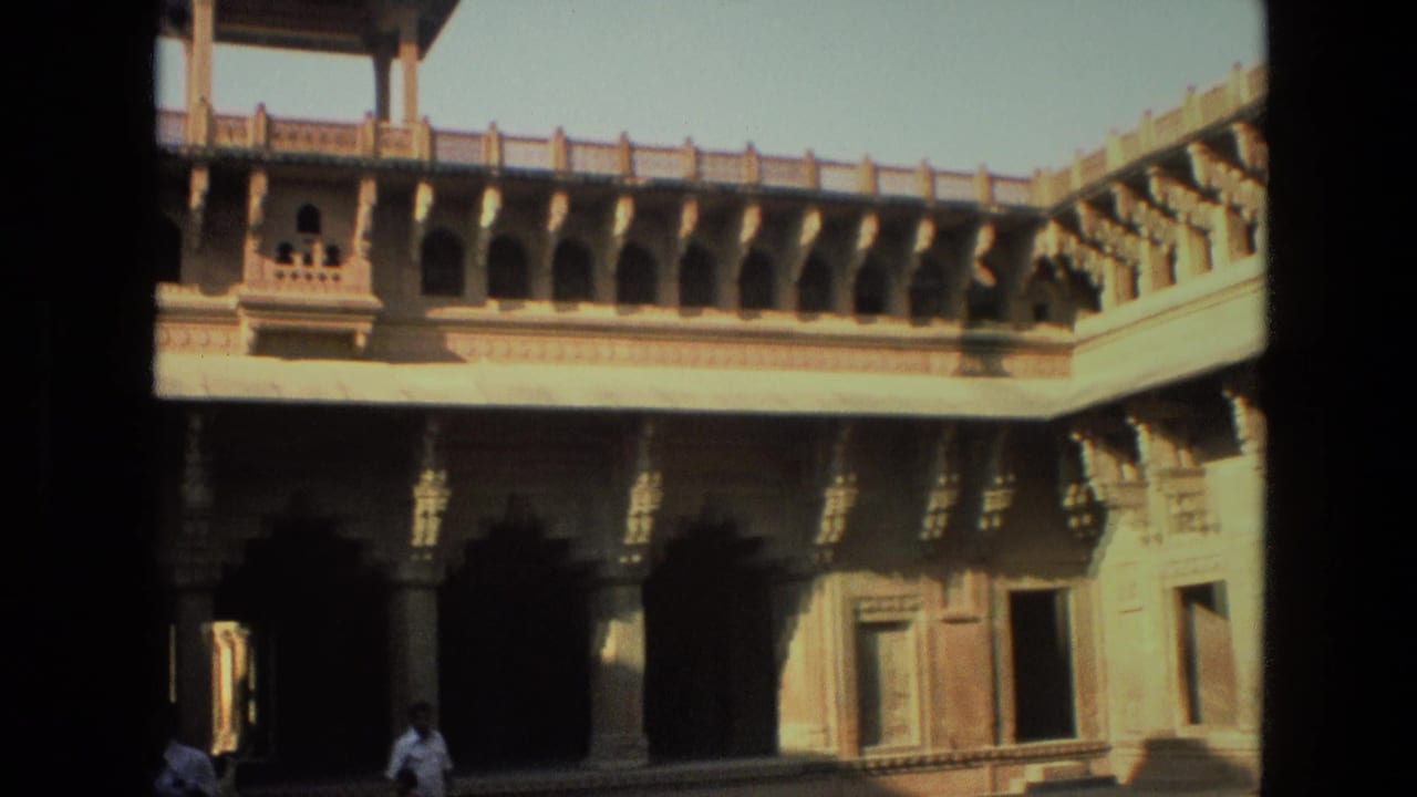 Sunlit stone courtyard with arched colonnade, carved pillars, and small visitors near the lower edge.