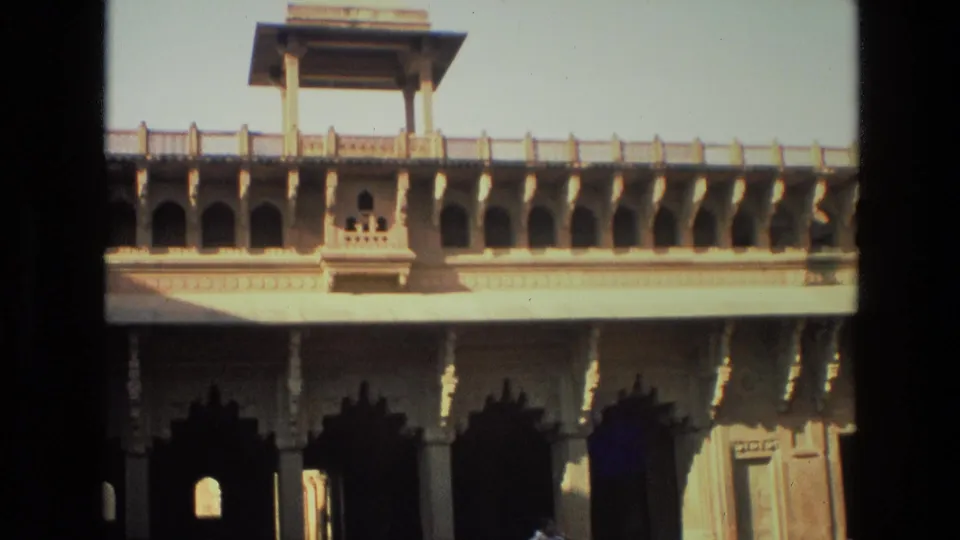 1974 India home-movie frame of an ornate stone arcade with an upper gallery, carved supports, and shadowed archways across the courtyard.