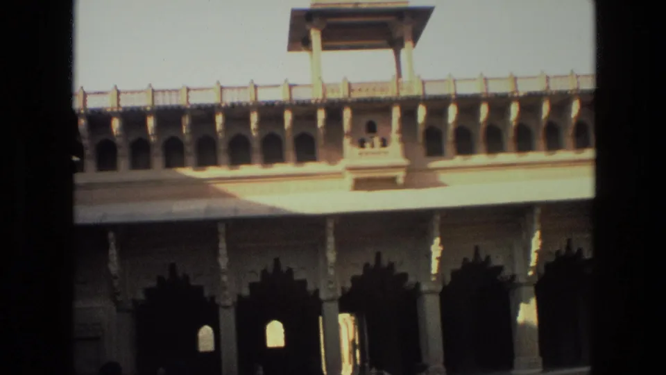 1974 8mm India footage showing repeated stone archways, carved columns, a projecting upper balcony, and strong sun across a courtyard facade.