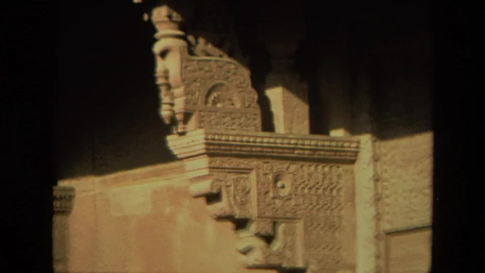 Close 1974 India travel-film view of a carved stone bracket or balcony support with a sculpted face, decorative relief, and warm side light.