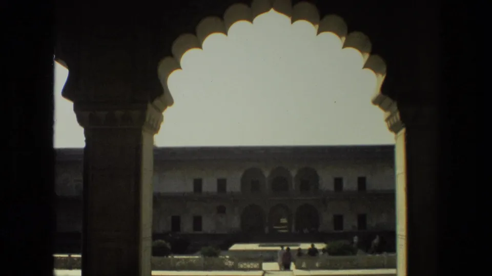 1974 India home-movie frame looking through a scalloped arch toward a courtyard facade with arched openings and distant visitors.