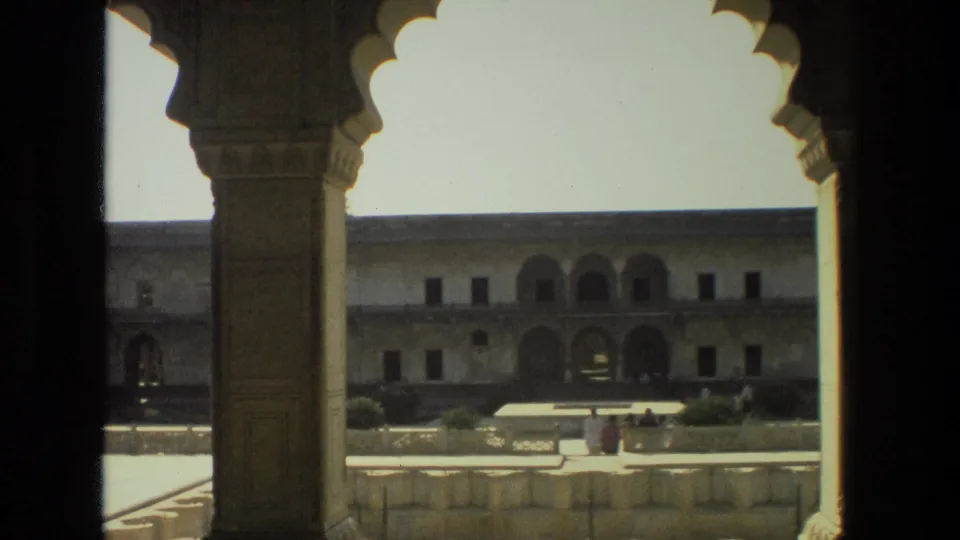 1974 India 8mm view of a sunlit courtyard facade, framed by a carved pillar and scalloped arch, with visitors near the lower edge.