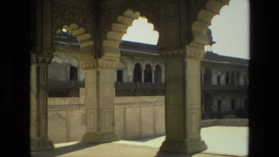 Final 8mm frame from a shaded arcade in India, looking through layered arches toward a palace or fort courtyard.