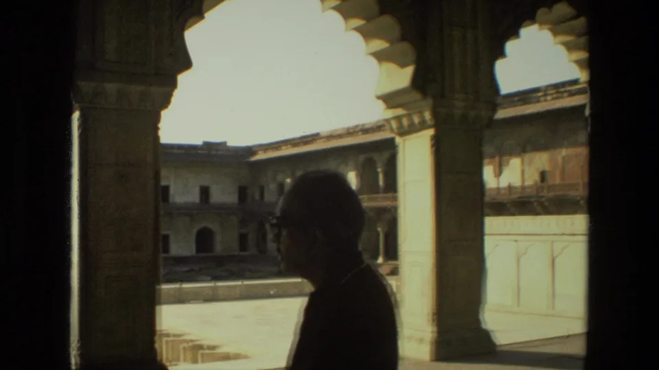 In 1974 India 8mm footage, an adult man in silhouette stands beneath carved arches looking toward a bright historic courtyard.