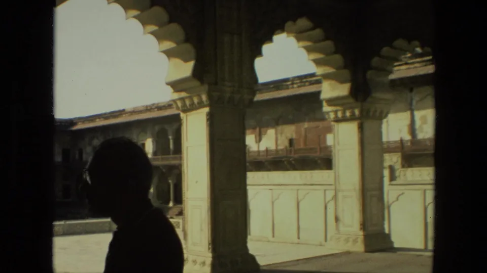 A silhouetted adult visitor is framed by scalloped arches and stone columns, with a bright Indian courtyard beyond.