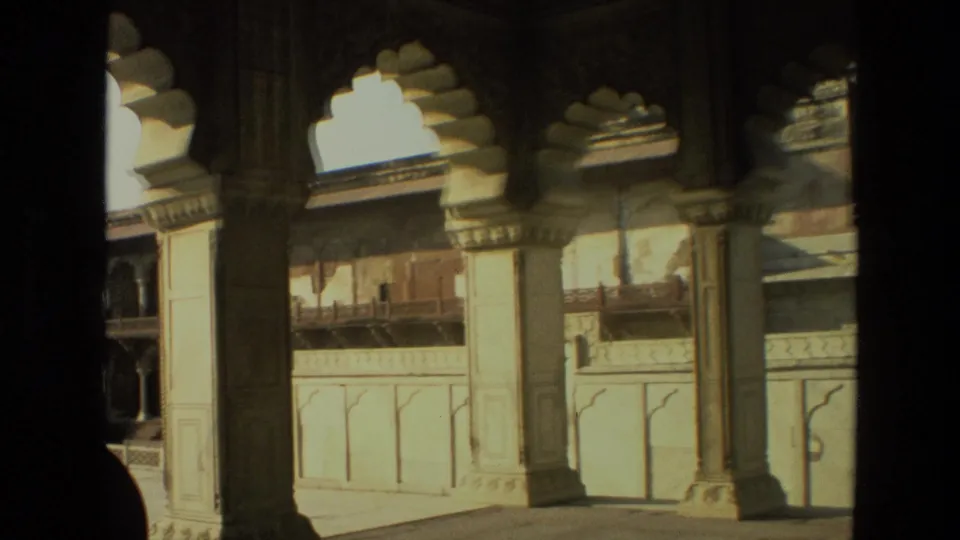 The camera shifts across carved pillars and arches, showing more of the sunlit courtyard and a partial silhouetted figure at the edge.