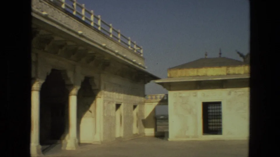 1974 India travel-film frame showing a marble colonnade, domed pavilion, barred window, and open courtyard under clear sky.