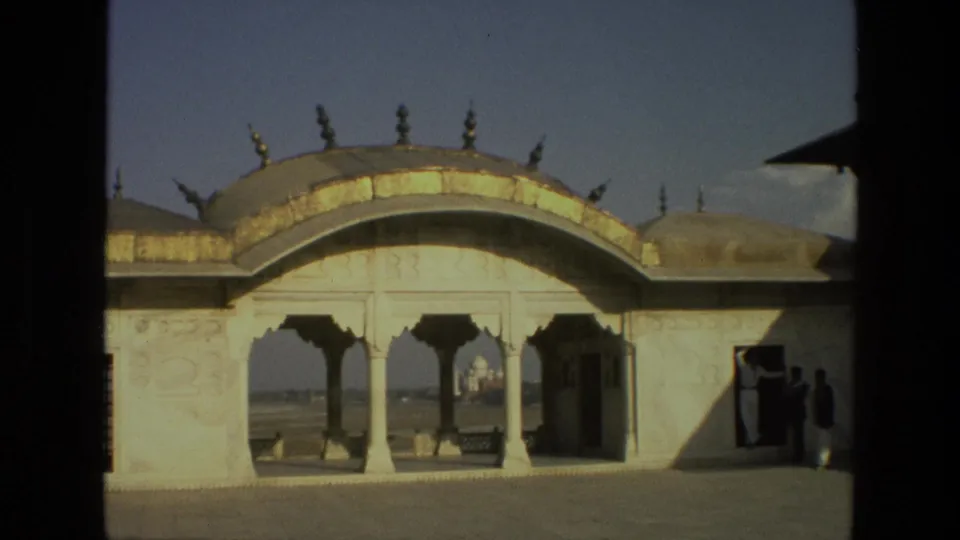 1974 India 8mm frame showing a sunlit arched pavilion, distant Taj Mahal, and small people near a shaded doorway.