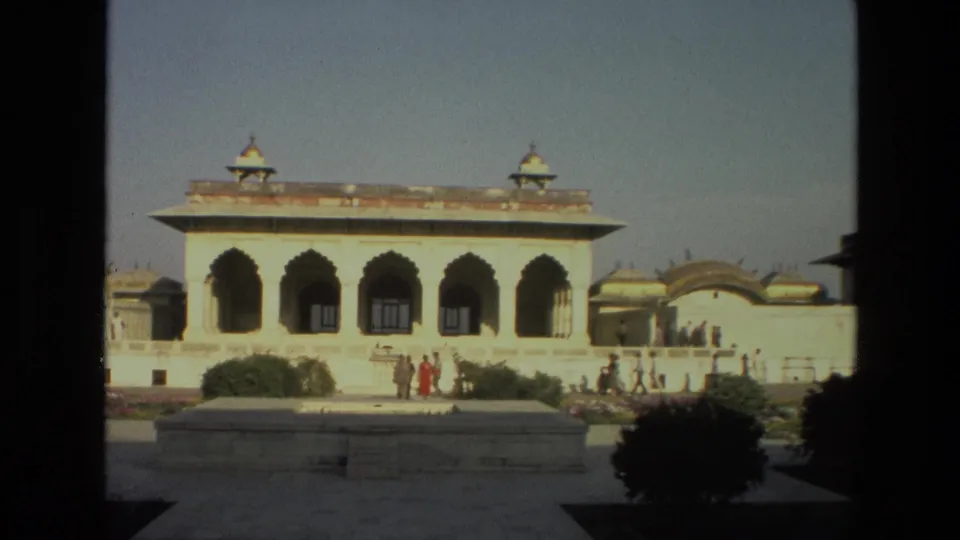 A historic Indian palace courtyard appears across a dark foreground frame, with arched openings, roof kiosks, and visitors outside.