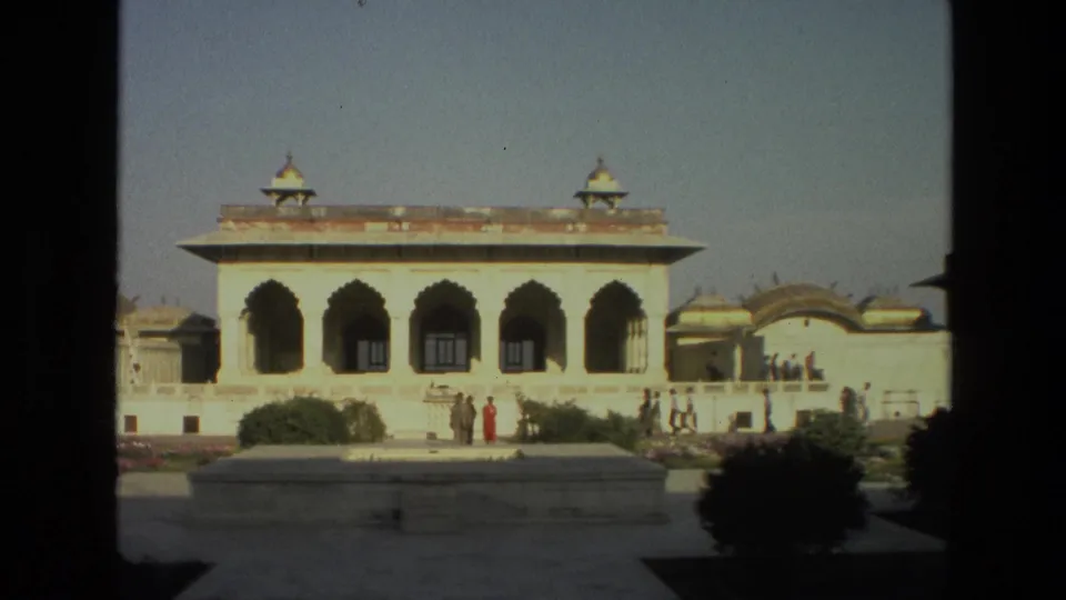 1974 8mm view of a pale arched palace facade in India, with small visitor groups and a red sari visible near garden planting.