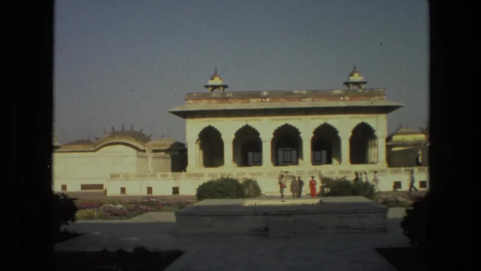 A sunlit 1974 India palace courtyard frame shows arched openings, a decorative wall, shrubs, and distant visitors in period clothing.