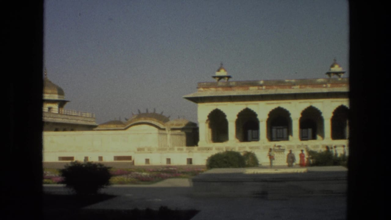 1974 India 8mm frame of a white arched palace facade, lawns, flowerbeds, and distant visitors partly framed by dark foreground edges.
