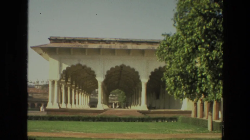 1974 India 8mm frame showing an open white arched colonnade, green lawn, clipped hedges, and a leafy tree within palace grounds.