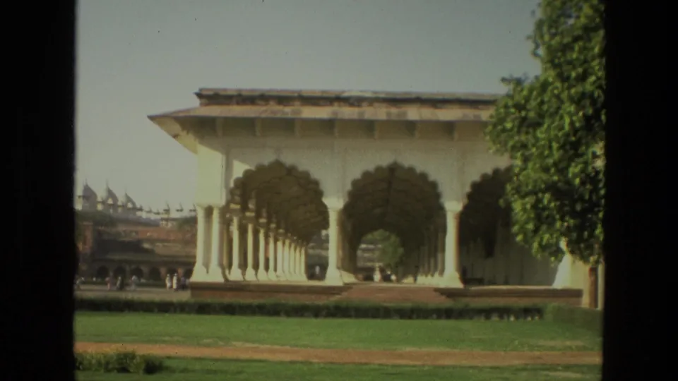 1974 India home-movie frame of a white arched colonnade, manicured lawn, low hedges, and a tree, with distant visitors barely visible beyond.