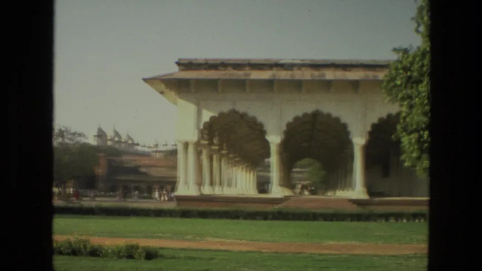 1974 India 8mm frame showing a pale arched colonnade, lawn, path, trees, and small visitors in the background.