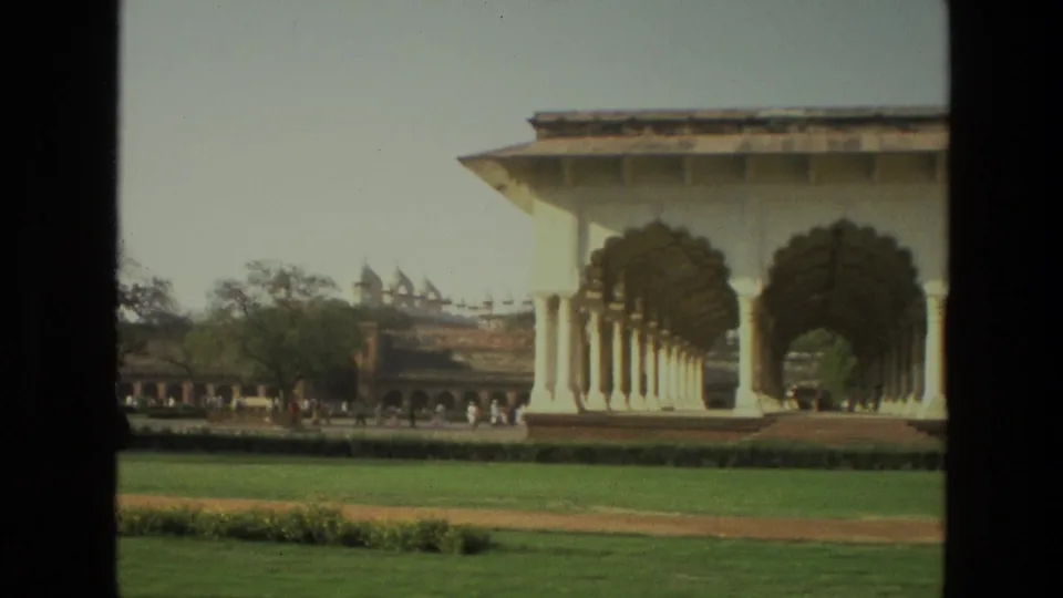 1974 India travel-film view of a landscaped courtyard with an arched pavilion, trees, distant wall, and small figures.