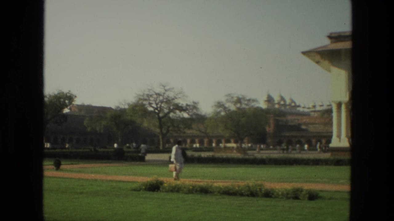 Garden lawn with arched building, distant domes, and a person in white walking near a path.