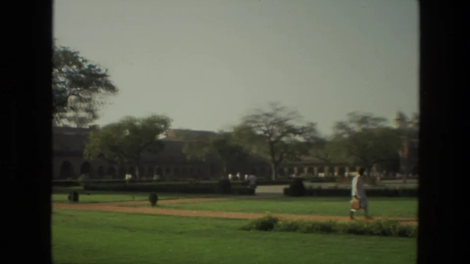 1974 India home-movie frame with a manicured garden, arched building line, trees, and a visitor walking near the right edge.