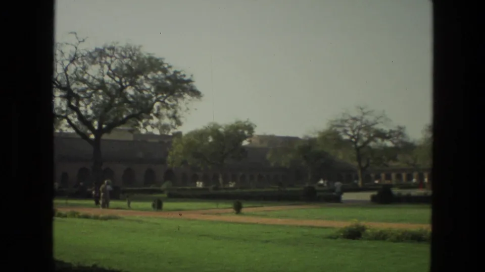 Wide 1974 India 8mm view of a formal garden with a large tree, long arched structure, paths, and distant visitors.