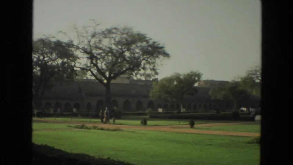 1974 India 8mm frame showing a formal lawn, path, arched background building, trees, and a small group of visitors walking.