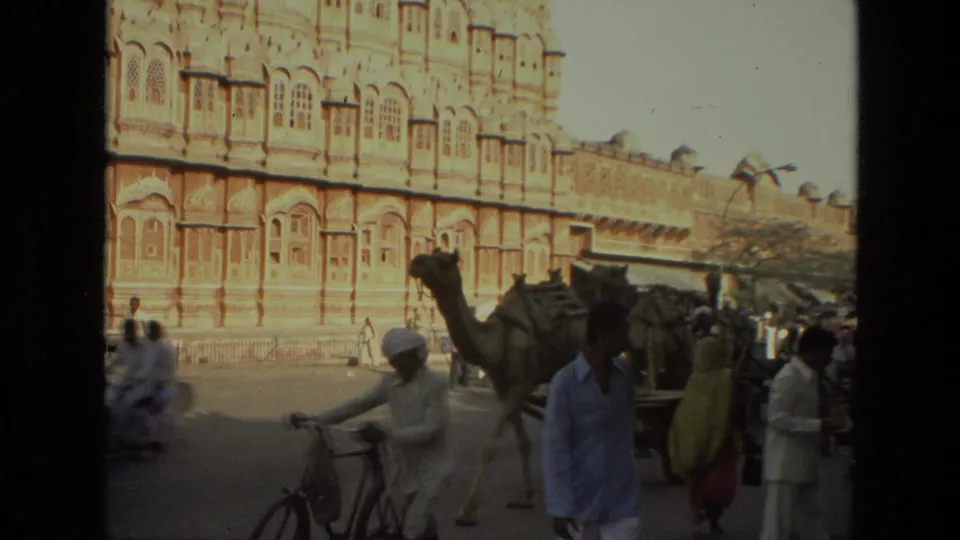 1974 India street view with a camel cart, pedestrians, bicycles, and the Hawa Mahal facade in the background.