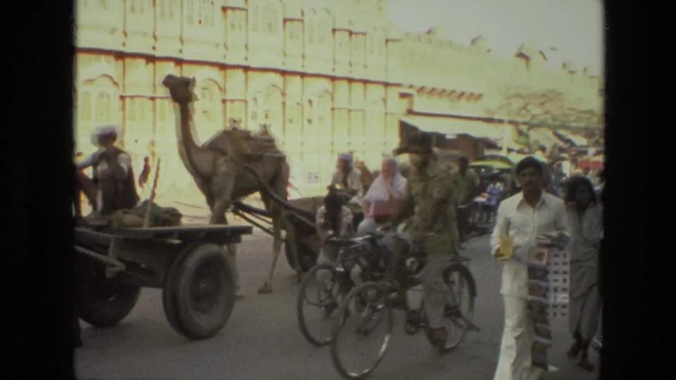 1974 India street view with a camel cart moving past cycle rickshaws, bicycles, pedestrians, and a pale ornate building facade.