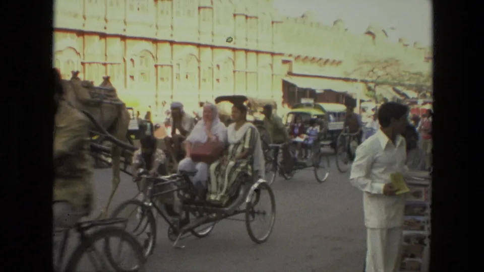 1974 India street footage with a cycle rickshaw in the foreground, a camel cart at left, pedestrians, bicycles, and market traffic.