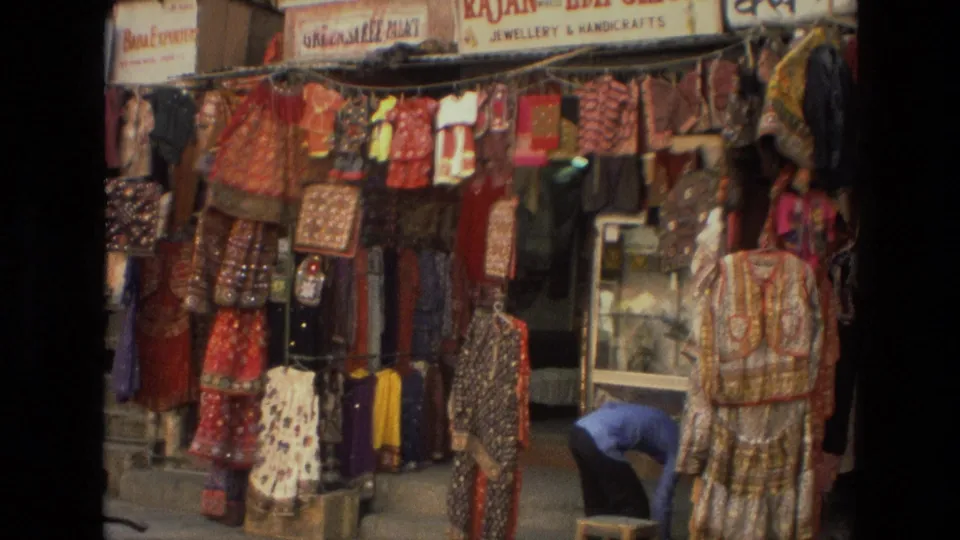 1974 India market shopfront showing colorful hanging textiles, garments, bags, shop signs, and a person working near the doorway.