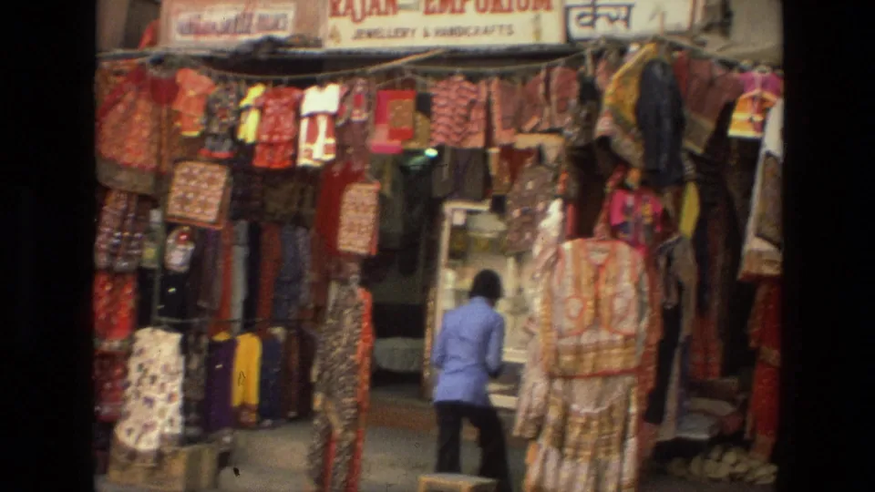 1974 India 8mm frame showing a woman in a blue top arranging merchandise at a dense traditional clothing stall.