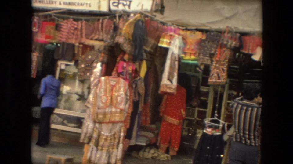 1974 India market frame with a woman arranging items beneath signs for an emporium and jewellery or handicrafts shop.