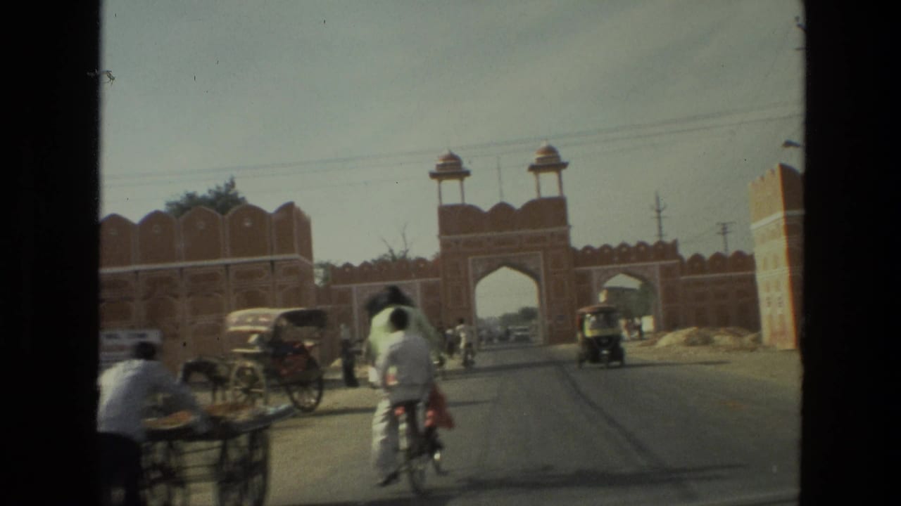 Street view toward a red sandstone gateway with cyclists, pedestrians, and auto-rickshaws.