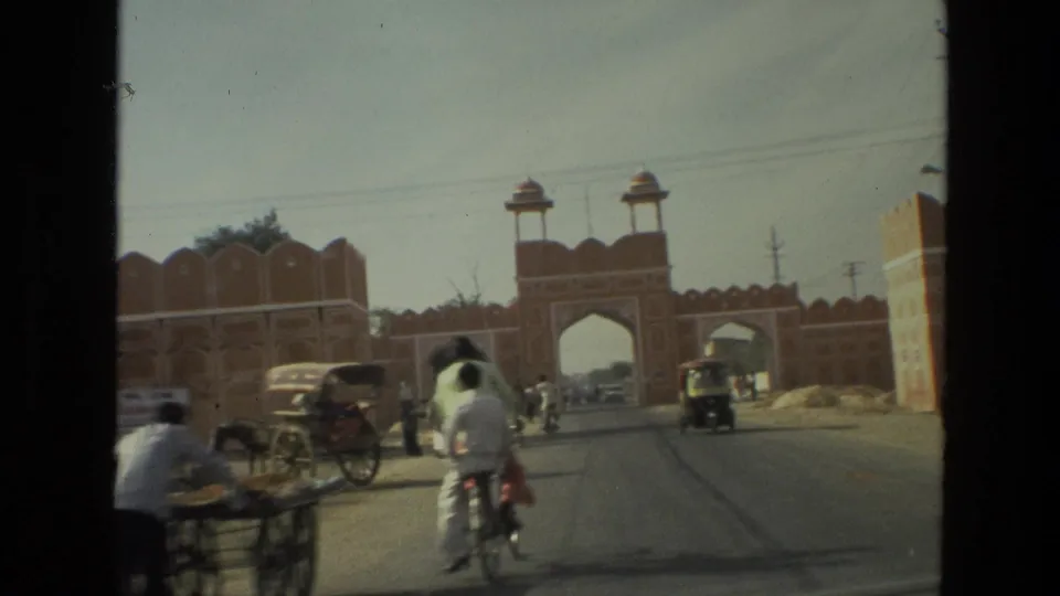 1974 India 8mm street scene approaching a historic-looking red sandstone gateway with cyclists, pedestrians, and auto-rickshaws.